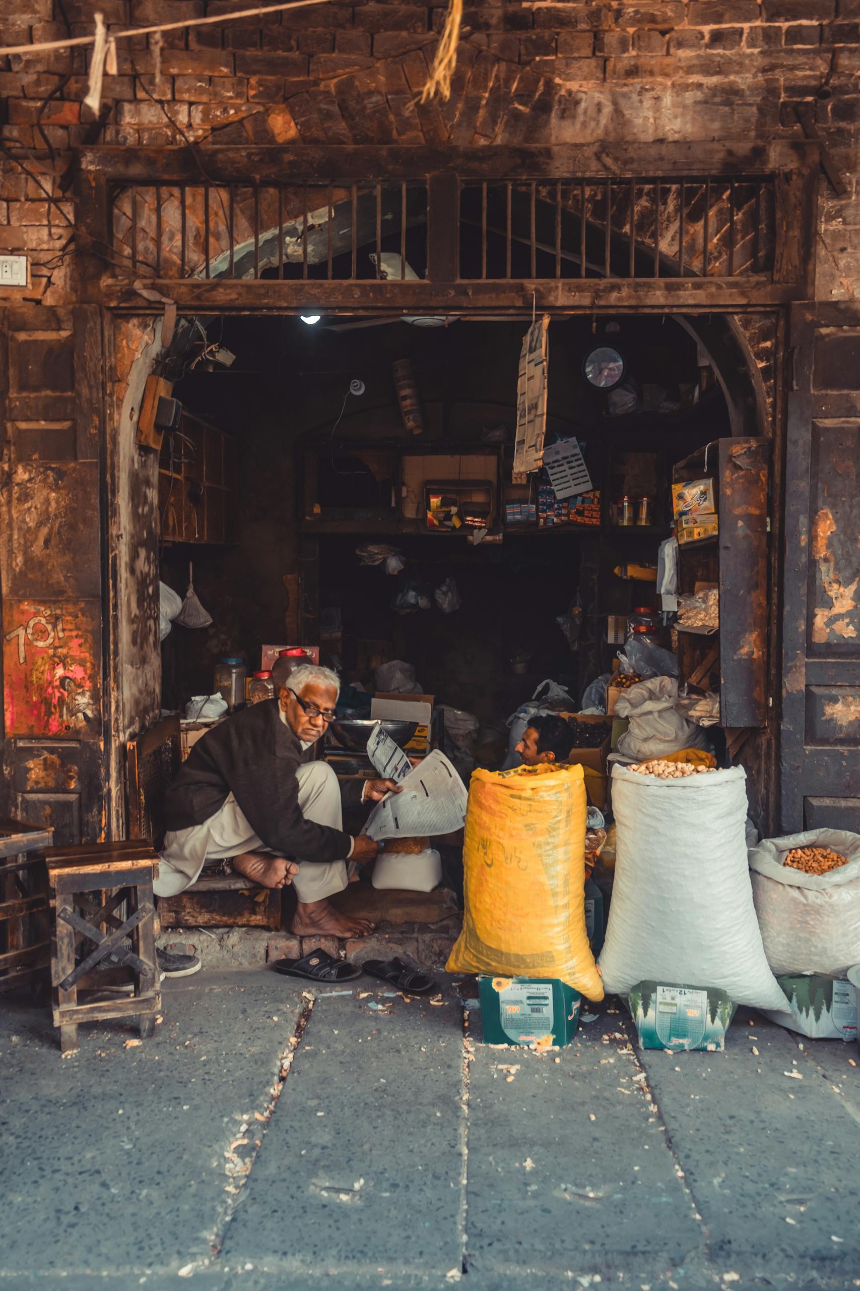 An elderly merchant in a traditional market setting in Pakistan, surrounded by bags of goods.