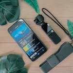Top view of a smartphone and gimbal surrounded by green leaves on a wooden surface.