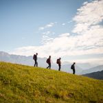Four hikers with backpacks walking downhill on a sunny day with mountains in the background.